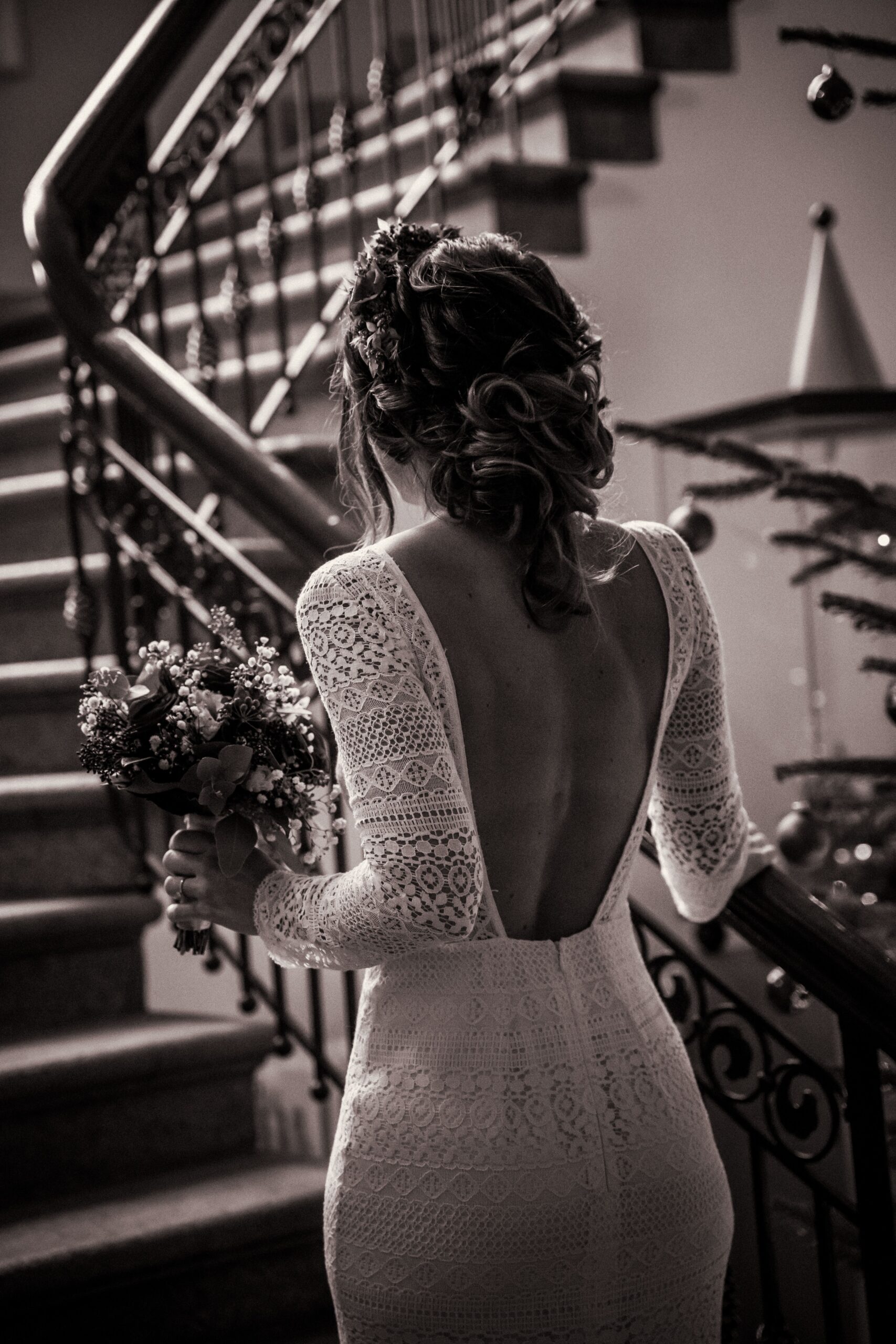 Mariée de dos tenant son bouquet de mariage dans la main gauche au pied d’un escalier